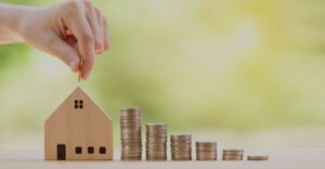 House-shaped coin bank with hand placing money inside it and stacks of coins to the right