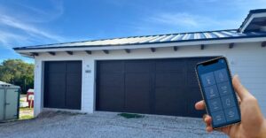 a smartphone displaying a smart home app, with the garage door highlighted, in front of a modern home's garage