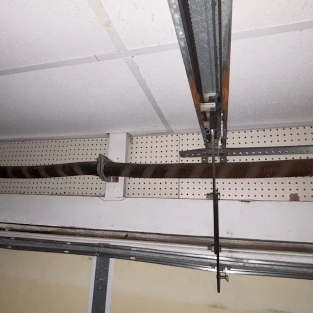 A detailed shot of a heavy-duty garage door torsion spring mounted on a metal shaft above the door, showing signs of wear requiring professional repair or replacement.
